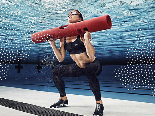 Woman doing squats underwater in a pool while holding a red foam exercise roller across her shoulders.